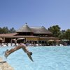 Persona tuffandosi in piscina, edificio con tetto di paglia sullo sfondo.