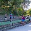 Persone giocano a bocce su un campo circondato da alberi; spettatori appoggiati alla recinzione.